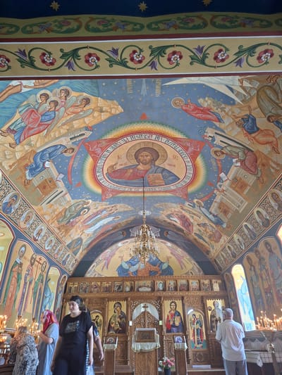 The interior of a church in Condrița, Moldova, features ornately painted ceilings and walls. The ceiling depicts religious scenes with figures like Jesus, angels, and other biblical characters rendered in vibrant colors. A large fresco of Jesus is centered in a circular motif on the ceiling, surrounded by a rainbow-colored halo and inscriptions. Below the ceiling, along the walls, are rows of painted icons of saints. The main focus in the foreground is the altar area, which includes an iconostasis, a screen or wall of icons, with several large, framed religious paintings. A chandelier hangs from the ceiling above the altar. Several individuals are present in the church. In the foreground, three women are walking or standing. Further back, a man is standing facing the altar, seemingly in prayer. Candles are lit on candelabra throughout the altar area, and floral arrangements are visible. The architecture includes arched windows and a coffered ceiling border with decorative floral patterns.