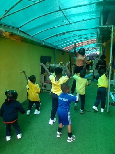 The image depicts a group of approximately eight to ten individuals, predominantly young children and one adult, engaged in a coordinated physical activity within a covered, semi-outdoor area in the city of Ajah, Nigeria. The environment features a translucent, corrugated turquoise roof supported by metal framing, a vibrant lime-green painted wall on the left, and a green artificial turf floor. Most children are dressed in yellow t-shirts and dark trousers or shorts, indicative of uniforms or activity attire. One young boy in the mid-foreground wears a blue t-shirt with "SAPPHIRE HOUSE" on the back and blue shorts, while a young girl in the left foreground wears a dark blue top and trousers with an orange stripe. An adult female, or older teenager, in an orange t-shirt and dark trousers, stands elevated towards the center-right with arms raised, appearing to lead the activity. All visible participants are performing similar movements with raised arms, suggesting an exercise, dance, or instructional session. Play equipment is partially visible in the background.