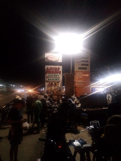 The image captures an outdoor night scene in the Sabo area of Abeokuta, Ogun State, Nigeria, focused on a commercial establishment. A prominent, bright overhead floodlight illuminates the central area. Two main vertical signs are visible: one features "LIVING PROB[S] EATERY BAKERY" in large green lettering on a white background, with images of baked goods below; the other, an orange vertical sign, lists services including "SARO SUPERMARKET," "EASY WAY," "SALON," and "RESTAURANT."

Approximately 15-20 individuals are present in the foreground and mid-ground. Most figures are indistinct due to backlighting and low light conditions, appearing as dark silhouettes. Individuals are observed standing, walking, and seated around small tables. One person in the mid-ground wears a light green top. In the left foreground, a person in dark attire holds a white object, possibly a bag.

The ground appears to be paved. Several small, dark tables and stools are arranged, particularly in the mid-ground and right foreground, suggesting an informal eating or vending area. Objects are visible on these tables, but specific details are obscured by shadow. Partial views of at least two motorcycles are discernible, one in the left foreground and another in the right foreground displaying a red light element. Distant vehicle headlights and other ambient light sources are visible in the background, indicating an active urban street or open space. Trash receptacles are situated near the base of the signs.