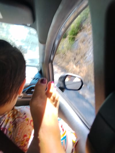 The image shows a person's foot resting on a car's dashboard, with the person's head visible in the background, looking forward. The foot has dark purple nail polish on the toenails. The scene appears to be inside a vehicle, with a view of the exterior through the window, suggesting the car is in motion. The exterior view shows a dry, hilly landscape with vegetation, consistent with the terrain found around Mattinata, Italy. The sunlight suggests it is daytime. The interior of the car is partially visible, including part of the dashboard and the car's side mirror reflecting the passing scenery.