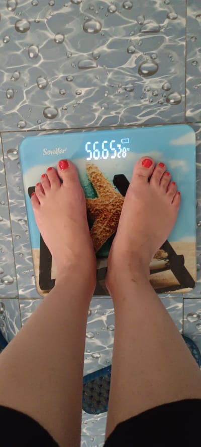 The image shows a person standing on a digital scale. The scale displays the weight as 56.65 kg and the temperature as 28 degrees Celsius. The person's feet, with red nail polish, are visible on the scale, which has a design of a starfish and sand. The scale is placed on a tiled floor with a water droplet pattern. The location of Baghdad, Uzbekistan is not depicted or suggested in this image.