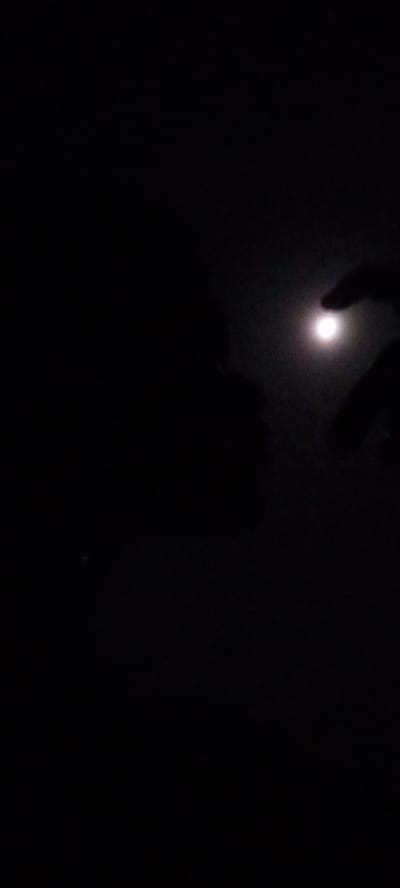 The image presents an extremely dark nighttime scene, dominated by deep black tones across most of the frame. In the upper right quadrant, a singular, bright, circular light source is visible, strongly resembling the moon, casting a soft glow around its perimeter.
In the immediate foreground on the right side, a dark, sharply defined silhouette of a human hand and forearm is captured, with fingers appearing outstretched towards the light source. To the left of the bright light, a larger, less distinct dark silhouette occupies the middle ground, suggesting the profile of a person's head and shoulders, seemingly facing or observing the light.
The overwhelming darkness indicates that the photo was taken at night in a very dimly lit or unlit environment. The activity appears to be an interaction with, or observation of, the moon or a similar celestial body, with a hand possibly attempting to "touch" or frame it. There are no other discernible objects, structures, or weather phenomena visible apart from the main subjects and the profound darkness. No text is visible within the image. While the provided location context is Ilogbo Eremi, Nigeria, there are no specific visual cues within the image itself to confirm this location.