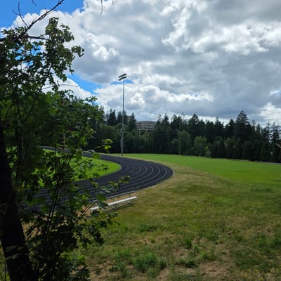 The image depicts a curving black running track set within a green grassy field. In the foreground, a large tree with dark bark and vibrant green leaves partially obscures the view of the track and field. A white bleacher with two rows of seats is visible alongside the track. Beyond the field, a dense line of evergreen trees forms a backdrop, with a multi-story building visible above the trees in the distance. A tall light pole with multiple fixtures stands near the track. The sky above is filled with dramatic, cloudy formations with patches of blue sky peeking through. The scene suggests an outdoor athletic facility, possibly a school or park, located in a suburban or semi-rural area. Based on the context provided, this location is in Portland, United States.