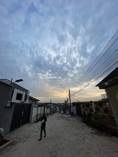The image shows a street in Isheri, Nigeria. The view is tilted to the side, capturing a cloudy sky overhead with power lines stretching across. Buildings line both sides of the street, with a two-story building on the right featuring plants on its balcony and a modern, gray building with a black metal gate on the left. A person is walking down the street on the right side. The road surface appears to be dirt and gravel.