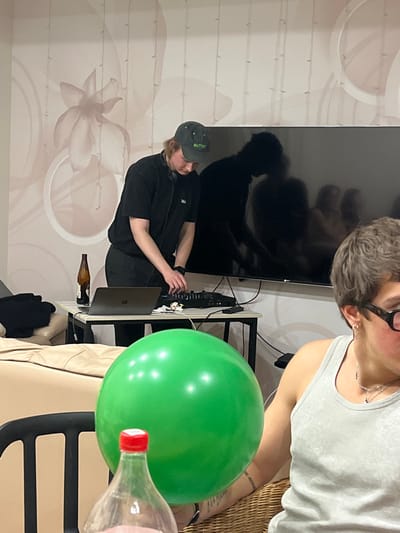 The scene depicts an indoor setting in Заозерное Снт, Russia. In the central portion of the image, a person with light-colored, shoulder-length hair, wearing a dark green baseball cap with "BUTTER" embroidered in light green, a black short-sleeved t-shirt, and headphones around their neck, stands leaning over a black rectangular table. This individual is actively manipulating a black DJ controller with both hands. To the left of the controller, a silver-colored laptop is open on the table, and a brown glass bottle is also present.
Behind the DJ setup, a large, flat-screen television is mounted on a light-colored wall. The television screen is dark and reflective, showing the silhouettes of several indistinct human figures. The wall itself features large, subtle, lighter-toned floral patterns and multiple vertical string-like decorative elements.
To the right, in the foreground, a second individual is partially visible, seated in a wicker chair. This person has short, dark, curly hair, wears dark-rimmed glasses, a grey ribbed tank top, multiple ear piercings, and a silver necklace. A large, bright green spherical balloon occupies the mid-foreground, partially obscuring this individual and a clear plastic bottle with a red cap, which is positioned closer to the viewer. The room is moderately lit.