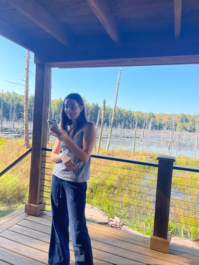 The image displays an outdoor scene from a wooden deck or viewing platform, overlooking a natural wetland or swamp area. While the specific city of Otego, United States, cannot be visually confirmed from the image, the environment is consistent with a rural, natural landscape.
A female subject, appearing to be in her twenties or thirties, stands centrally on the deck, facing slightly towards the viewer. She has long, dark hair and is wearing a light-colored, possibly plaid or gingham, sleeveless top with a V-neck, and dark blue, wide-leg trousers with a visible drawstring at the waist. She holds a silver-colored smartphone in her left hand, positioned at chest level, and her right hand rests near her stomach. Her expression is a slight smile directed towards the camera.
The deck features light-colored wooden plank flooring and dark-stained wooden support posts. A black metal railing with horizontal tension cables separates the deck from the natural environment. Beyond the railing, a body of water is visible, partially obscured by green and golden-brown marsh vegetation. Numerous skeletal, light-colored tree trunks protrude vertically from the water, characteristic of a flooded forest or beaver pond. In the background, a dense forest displays early autumnal foliage with green, yellow, and orange hues. The sky is clear and bright blue, indicating sunny weather.