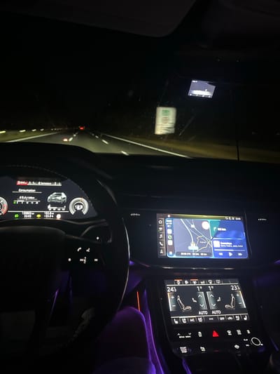 The image displays the interior of a modern vehicle from a driver's or front passenger's perspective, traveling at night on a multi-lane road. The car's digital instrument cluster shows a speed of 123 km/h and a current fuel consumption of 9.4 l/100km. The total odometer reading is 130,863 km, and the outside temperature is 18.3°C. The dashboard clock indicates 22:03.

To the right of the steering wheel, a large touchscreen infotainment system displays a navigation map. The map shows a route with a destination or point of interest labeled "Αιγίνιο" (Aiginio) in Greece. The system indicates an estimated time of arrival (ETA) of 22:50 and a remaining distance of 60 km. Simultaneously, a media player widget on the screen displays "Somedays" by "Sonny Fodera, Jazzy, D.O.D".

Below the infotainment screen, another touchscreen panel controls the climate system. It shows dual-zone settings of 24.5°C for the left side and 23.5°C for the right side, along with controls for heated/ventilated seats and air conditioning. Additional car controls, including "drive select", "START ENGINE STOP", and the hazard lights button, are visible on the console. Purple ambient lighting is present in the lower cabin area.

Through the windshield, the dark road ahead is illuminated by the car's headlights, revealing white lane markings. Red taillights of a distant vehicle are visible on the road. A small electronic device, possibly a dashcam or radar detector, is mounted on the windshield near the top. A white rectangular object with green markings, resembling a toll pass or permit, is also affixed to the windshield. No individuals are explicitly visible within the frame.