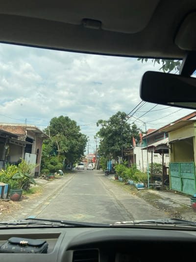 The image depicts a residential street scene in Ketegan, Indonesia, viewed through the windshield of a vehicle. The sky overhead is partly cloudy, with a mix of white and grey clouds. The foreground shows the dashboard and lower portion of the windshield, with a dark, rectangular object (possibly a wallet or small bag) resting on the dashboard. The vehicle's rearview mirror is visible in the upper right quadrant, partially obscuring the sky and distant trees.
The street is a narrow, paved road, possibly made of patterned concrete or cobblestones, extending into the distance. It is bordered on both sides by unpaved or partially paved shoulders containing dirt and vegetation. Residential buildings line both sides of the street. On the left, a brick and light-colored building with a sloping roof is visible, partially obscured by lush green plants in decorative pots. On the right, houses with various facades are present, including one with a light yellow exterior and a green metal gate, and another further down displaying a red and white flag, likely the Indonesian national flag.
Abundant green foliage from trees and potted plants lines the street on both sides. Overhead, a network of black utility wires is visible, strung between multiple utility poles. In the mid-distance, two light-colored cars, possibly sedans or small vans, are visible on the road, oriented away from the observer. A dark-colored scooter or motorbike is parked on the right side of the road, partially obscured by plants. No individuals are visible in the scene. The overall impression is a quiet, residential neighborhood during daylight hours.