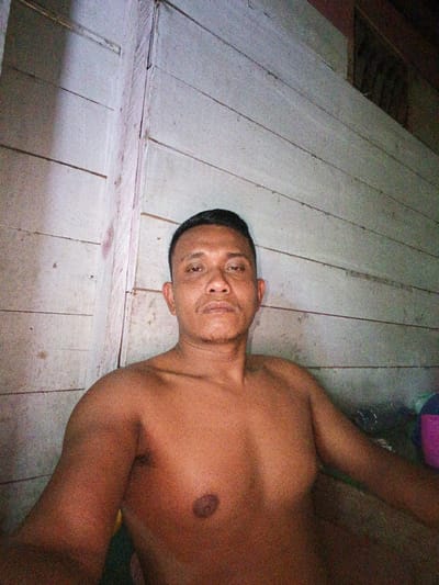 A bare-chested man of Asian descent is seated in front of a light-colored, horizontally paneled wooden wall. He has short, dark hair and is looking directly at the camera with a neutral expression. His skin tone is brown. The background wall appears to be made of planks, with a vertical wooden beam visible on the left side of the image. The lighting is dim, casting shadows and creating a grainy texture throughout the image. To the right of the man, out of focus, are some objects including a purple container. The scene appears to be indoors, possibly in a rustic or unfinished dwelling.