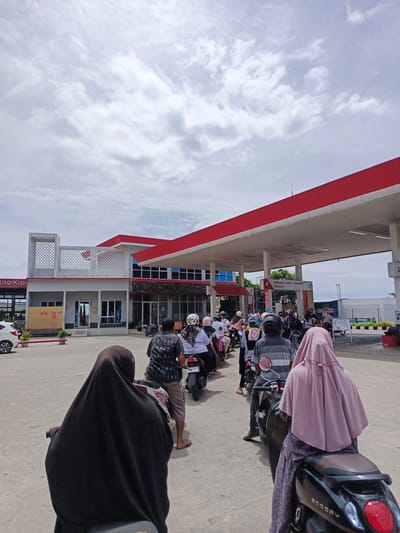 This is a wide-angle, eye-level shot of a queue of motorcyclists at a gas station in Cotbadatunong, Indonesia. The gas station has a red and white canopy supported by white pillars. In the background, there is a building with white and grey elements, and some signs. The sky is overcast with white clouds. The foreground features a line of motorcycles with riders, most wearing helmets. Two individuals in the immediate foreground are riding motorcycles and are partially visible from behind, one wearing a black hijab and the other a pink hijab. The motorcyclists are lined up waiting to refuel. The ground is paved concrete.
