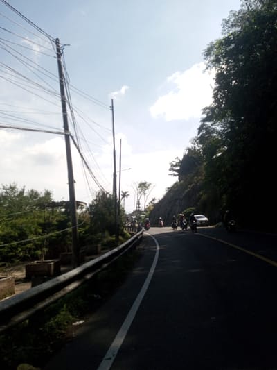 The image depicts a winding asphalt road in Ban Phonhxavanh, Laos, under a bright, partly cloudy sky. The road, featuring a white solid line marking its left edge, curves rightward into the distance. A dark metal guardrail runs along the left side of the road, behind which are several wooden utility poles densely crisscrossed with numerous electrical and communication wires. Beyond the poles, lush green vegetation, including trees and shrubs, is visible, along with several concrete planters. On the right side of the road, a steep, rocky hillside is covered with dense foliage and large trees, casting significant shadows onto the road and parts of the hill itself.

Multiple individuals are visible riding motorcycles or scooters on the road, primarily moving away from the viewer, suggesting traffic flow. At least one white sedan is parked on the right shoulder of the road further down the curve. The scene is illuminated by strong sunlight, creating bright highlights and deep shadows.