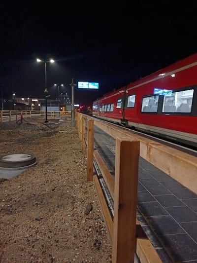 At night, a red passenger train is stopped alongside a station platform with wooden railings. Streetlights illuminate the platform and surrounding gravel area. The train's lit windows reveal interior seating. Signage is visible above the platform, and other lights can be seen in the distance. A manhole cover is in the foreground, adding to the scene's setting.