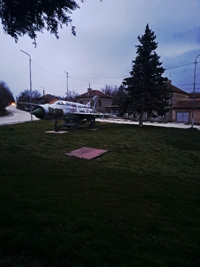 The image depicts an outdoor scene in Kamenets, Bulgaria, under an overcast, muted blue-grey sky suggestive of dusk or dawn. A decommissioned MiG-21 fighter jet, featuring a weathered camouflage pattern of greens, browns, and light grey with a prominent green nose cone, is centrally positioned on a maintained grassy area. The aircraft is mounted on a pedestal, oriented with its nose pointing towards the left foreground.

In the background, a paved road curves gently to the left, bordered by bare deciduous trees and sparse winter vegetation. Along the road and behind the grassy area, several single-story residential or small commercial buildings with tiled roofs and light-colored facades are visible. Faint orange vehicle lights are discernible in the distance on the far left section of the road.

To the right of the aircraft, a tall, dark green coniferous tree stands prominently on the grassy expanse. Two modern streetlights with vertical poles and rectangular LED fixtures are present: one behind the jet on the left, and another near the coniferous tree on the right. Overhead utility wires traverse the mid-ground. In the lower center of the foreground, a flat, rectangular concrete or metal slab, possibly an access panel, is embedded in the grass. Dark foliage from an unseen tree partially frames the upper left corner of the scene. No individuals are present, and no explicit actions or interactions are depicted.