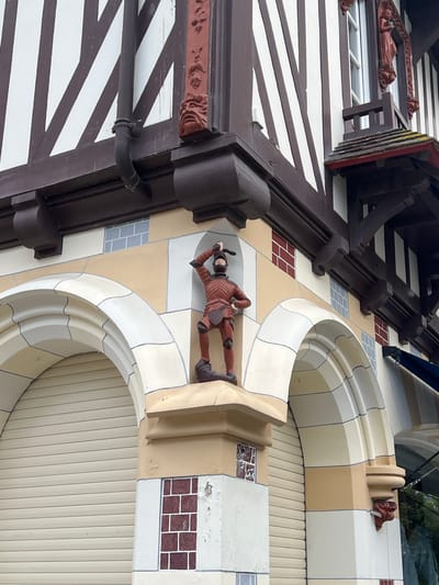The image shows the exterior of a building in Deauville, France, with distinctive half-timbered architecture and decorative elements. The building features dark brown beams against a cream-colored wall, creating a geometric pattern. A drainpipe is visible along the side of the building.
On the lower section of the building, there are large arched openings, one of which has a rolling shutter. A statue of a knight in armor stands on a ledge above one of the arches. The statue is reddish-brown and appears to be made of wood or a similar material. The knight is depicted in a dynamic pose, holding a sword above his head. The facade is decorated with painted brickwork and geometric patterns. There are also grotesque masks adorning the building's architectural details, such as on the corners of the upper story and near the arches.
The overall impression is of a historical or themed building, possibly a shop or restaurant, with a whimsical and ornate design. The sky is overcast, suggesting a typical day in Normandy.