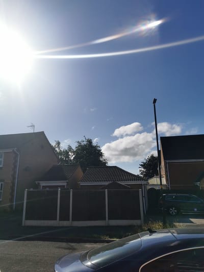 The image depicts a residential street scene in Ripon, United Kingdom, under a bright, sunny sky with scattered clouds. The sun is high, causing lens flare across the upper portion of the frame, creating streaks of light and rainbow-like refractions.
In the foreground, the roof of a dark blue car is visible, reflecting the sunlight. To the right, a black station wagon is parked on the street. Behind the cars and the street is a dark fence with concrete posts, separating the properties.
Further back, several houses with tiled roofs are visible. One house on the left is made of brick, with white trim around the windows and a TV antenna on its roof. Adjacent to it are smaller structures, possibly garages or single-story extensions, also with tiled roofs. A tall, slender street lamp stands to the right of center, its light fixture at the top. More brick houses with dark roofs are visible in the background. Trees, some with dark foliage, are interspersed between the houses. The environment appears to be a typical suburban street.