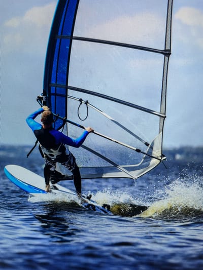 A man windsurfing in the waters off Bacong, Philippines. The man, wearing a wetsuit, is standing on the windsurfing board, holding onto the sail. Splashes of water are visible where the board cuts through the water. The environment is an expanse of water under a bright sky.
