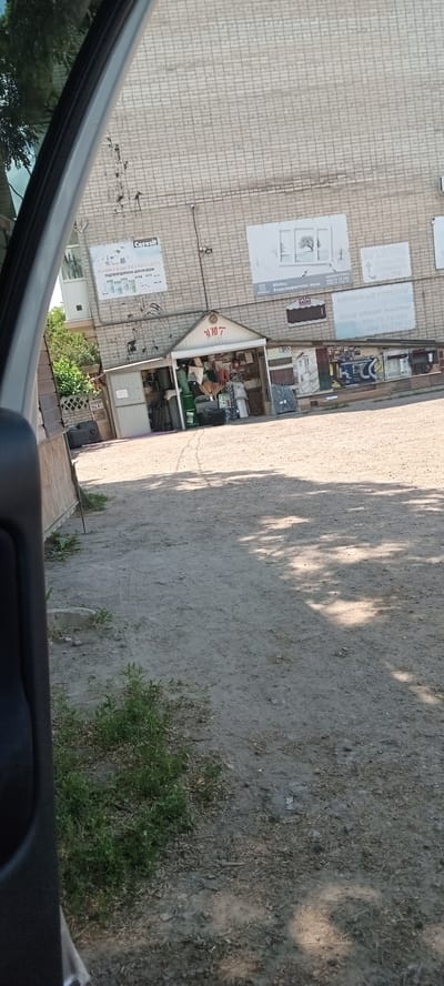 A low-angle, wide shot captures a small roadside market stall in Valky, Ukraine, on a sunny day. The stall, a simple, open-fronted structure with a white roof and a sign bearing the Cyrillic letters "УПО7" (UPO7), displays a variety of goods including tires, construction materials, and what appear to be cleaning supplies. Behind the stall, the brick wall of a larger building is visible, adorned with several advertisements. One large sign features the "Ceresit" logo and text in Ukrainian, promoting building materials. Another advertisement displays a picture of a tree in a frame, with text mentioning "магазин «Укрта»" (Ukta store) and contact information. To the left of the stall, a gray metal storage unit stands against the building. The ground in front of the stall is dirt, with some scattered debris and tire tracks. Lush green vegetation grows along the left side of the frame, near a low fence. The overall scene suggests a commercial area on the outskirts of the city, where a small business operates.