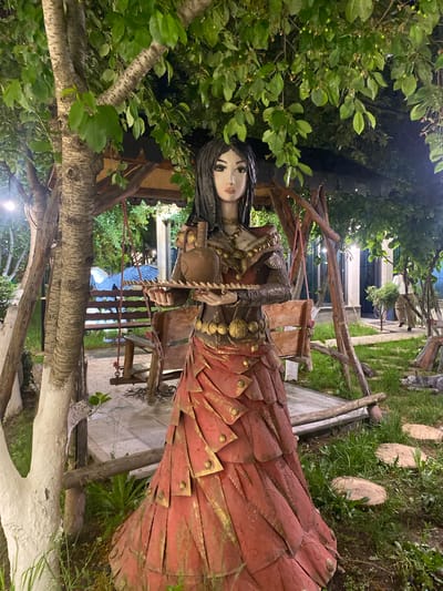 The image captures a sculpture in Verin Ptghni, Armenia. The sculpture depicts a female figure in a red dress adorned with gold details. The sculpture is surrounded by greenery, including trees and grass. The location appears to be a garden or outdoor space, with wooden structures and stepping stones visible in the background.