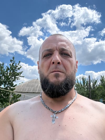 The media file is a close-up, low-angle selfie of a man taken outdoors. The man, who appears to be middle-aged with a receding hairline, short beard, and a stern expression, is wearing a silver chain necklace with a cross pendant. His bare chest is visible, and his skin has several moles.

The background consists of a bright blue sky with fluffy white clouds, indicating fair weather and daytime. Green trees and a portion of a building with a corrugated roof are also visible, suggesting a rural or suburban setting. The orientation of the image is rotated, with the man's head appearing at the top.

The scene appears to be a casual moment, possibly a self-portrait taken during a sunny day. There is no visible text in the image. The location context provided is Zhirnov, Russia.