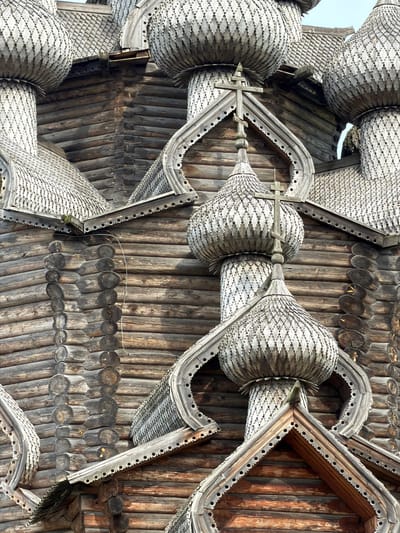 This image is a close-up view of the traditional wooden architecture of a Russian Orthodox church located in Nevskii Parkleskhoz, Russia. The focus is on the intricately carved wooden domes and roof structures, which are characteristic of Russian wooden church construction. The domes are covered with wooden shingles arranged in a scale-like pattern, and each is topped with a cross. Decorative wooden trim with carved patterns adorns the edges of the roofs and around the domes. The walls of the church are constructed from dark, weathered logs, showing the texture and grain of the wood. The sky is visible in the upper part of the image, suggesting an outdoor setting. There are no people visible in the frame. The overall impression is one of historical craftsmanship and religious architecture.