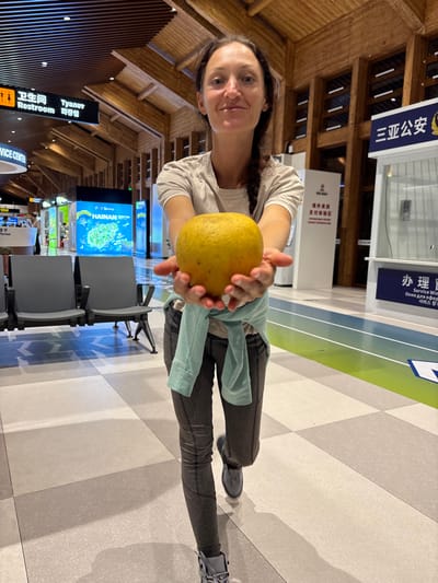 The image captures a female individual holding a large yellow citrus fruit within Sanya Phoenix International Airport (SYX) in Tianya District, Sanya, Hainan, China. The woman, positioned centrally and facing the camera, has dark hair, wears a light short-sleeved top, dark grey patterned leggings, and light grey athletic shoes, with a light blue shirt tied around her waist. She extends both hands forward, presenting the fruit towards the viewer, and exhibits a slight smile.

The environment is a modern airport terminal featuring a distinctive wooden slatted ceiling and a light-colored tiled floor composed of grey and beige squares. Bright artificial lighting illuminates the scene. In the background to the left, airport seating (dark grey, bench-style) is visible, along with a black "Restroom" sign displaying text in Chinese, English, and Korean. Further back, illuminated display boards include one with "HAINAN" and an underwater image. To the right in the background, white service kiosks are present, one featuring a blue sign with "三亚公安" (Sanya Public Security) in Chinese and partially visible English text. Another sign for "HILTON" is also discernible.