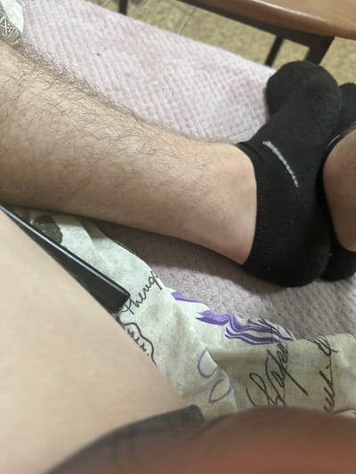 In Corlăteni, Moldova, a close-up, eye-level shot shows a person's legs resting on a pink textured surface, likely a bedspread. The legs are covered in dark socks, and one leg is hairy and bare. A black object, possibly a pen or stylus, lies on a patterned fabric. The scene is indoors and appears to be a private, relaxed setting.