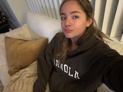 A young woman is sitting on a white couch with a brown hooded sweatshirt with “MANOLA” printed on it. She is looking at the camera with a slight pout. Behind her, there are closed white blinds. There are two pillows and a textured throw on the couch next to her. There is a side table with dark objects sitting on it behind the couch. She has light brown hair. She is wearing small gold hoop earrings.