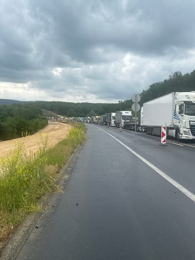 The image depicts a traffic jam on a road in Tatabánya, Hungary. A line of white tractor-trailer trucks are stopped on the right side of the road. A white line is painted on the asphalt. The sky is overcast with dark clouds. Vegetation grows along the side of the road and up a hill. The location appears to be rural.