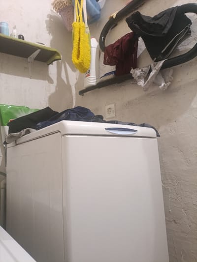 The image shows a cluttered laundry room or utility space. A white top-loading washing machine is prominently featured in the foreground, with some dark clothing draped over its top. To the left of the washing machine, a portion of a white bathtub or sink is visible.
Above the washing machine, a narrow shelf holds a few items, including a bottle and a black object that could be a cap or lid. Further up and to the right, a black pipe rack is mounted on the wall, with several items of clothing, including dark shorts and a burgundy shirt, hanging from it. A yellow, mesh-like bath pouf and a knitted item are also hanging on the wall in the upper left. A white plastic bottle with a red label is visible on the shelf to the right of the yellow pouf. A blue-handled brush is resting on the pipe rack. A white electrical socket is visible on the wall in the background. The walls appear to be roughly textured and off-white.
The scene suggests a lived-in and functional space, likely in a residential setting. The presence of laundry and cleaning supplies indicates ongoing domestic chores. The lighting appears to be artificial, suggesting it could be any time of day. There is no visible text in the image.