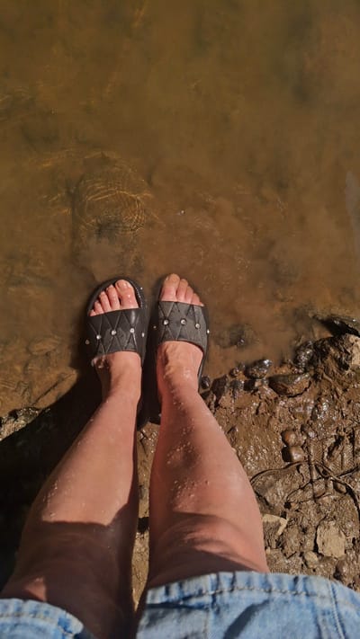 The image shows a top-down view of a person's legs and feet standing in murky water next to a muddy bank. The water is brown and appears shallow, with some ripples on the surface. The ground on the bank is wet and muddy, with a few rocks and debris visible. The person is wearing black slide sandals with decorative rhinestones and denim shorts. The scene appears to be outdoors, possibly near a body of water like a river or pond. There is no specific indication of the location within Дзяржынскі сельскі Савет, Belarus.