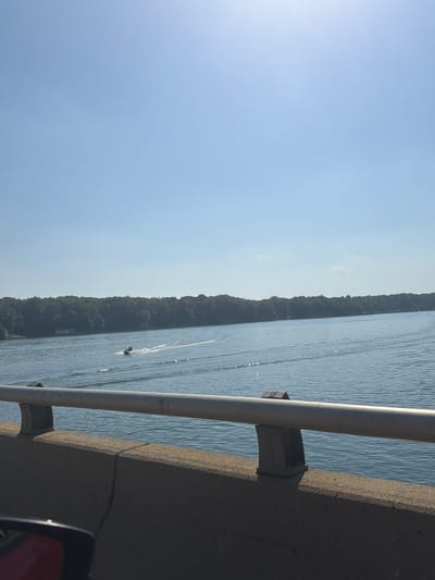 A wide-angle shot captures a serene lake under a clear, bright blue sky. In the distance, a dense line of green trees forms the shoreline. Closer to the viewer, a jet ski with two people is actively moving across the water, leaving a white wake behind it. The foreground features a concrete barrier with a metal railing, suggesting the photo was taken from a bridge or elevated roadway. The sun is high in the sky, casting a bright light that reflects off the water's surface, creating shimmering patterns. The overall scene depicts a recreational activity on a calm body of water on a sunny day.