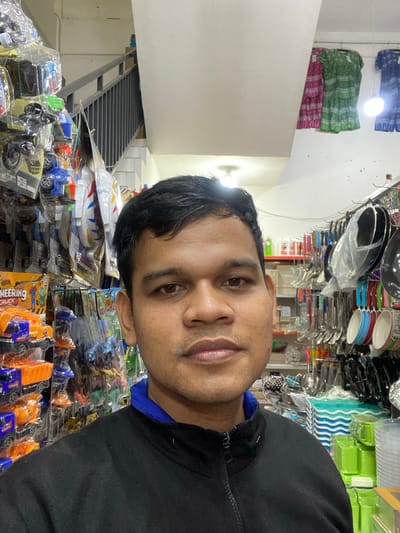 The media file is a selfie taken in what appears to be a retail store, possibly in Lamteumen Barat, Indonesia. The subject is a man with dark hair and brown eyes, facing the camera. He is wearing a black hooded jacket with a blue undershirt. His expression is neutral.
The background is filled with shelves of various products. On the left side of the image, stacks of what appear to be folded textiles in blue, green, and pink patterns are visible. Above these, a white wall is present, and to the right, a metal staircase is visible.
Further to the right, the shelves are packed with a variety of items. Some are clear plastic containers, others are colored kitchenware like plates and bowls in red and blue. There are also many toys, predominantly toy trucks and construction vehicles, some packaged in plastic. The overall impression is of a crowded retail environment selling household goods and toys.
The lighting in the scene seems to be artificial, with bright light sources illuminating the products. There is no indication of the time of day or weather conditions. The presence of a wide array of consumer goods suggests the setting is a shop or market.
Visible text includes "ENGINEERING TRUCK" on the packaging of some of the toys. The orientation of the image is rotated.