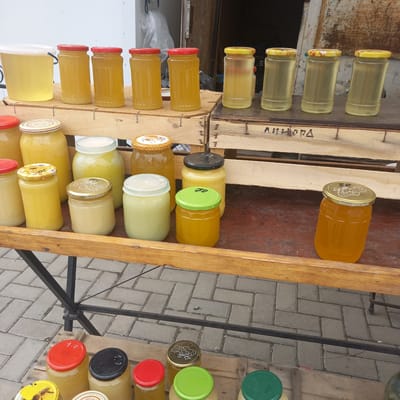 The image depicts an outdoor market stall in Briceni, Moldova, displaying an assortment of honey jars. The stall features wooden crates and shelves, upon which various sizes of glass jars filled with honey are arranged. The honey varies in color from a light yellow to a deep amber. Some jars have decorative lids, while others have plain colored caps, some of which are marked with prices or lot numbers. In the foreground, more jars of honey are visible on a lower wooden surface, some with brightly colored lids. The ground is paved with interlocking gray bricks. The overall scene suggests a local vendor selling honey.
