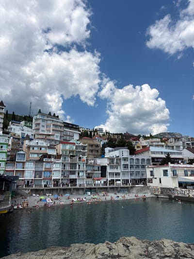The image depicts a coastal town with multi-story buildings constructed on a hillside overlooking the sea. The buildings are a mix of modern and older architecture, with many featuring balconies and colorful facades. A beach is situated at the base of the buildings, populated by numerous individuals sunbathing and swimming. The sea is calm and dark blue. The sky above is partly cloudy with fluffy white clouds against a bright blue background. The scene appears to be a popular tourist destination, likely during the summer season. The location is identified as Kiparisnoe, Ukraine.
