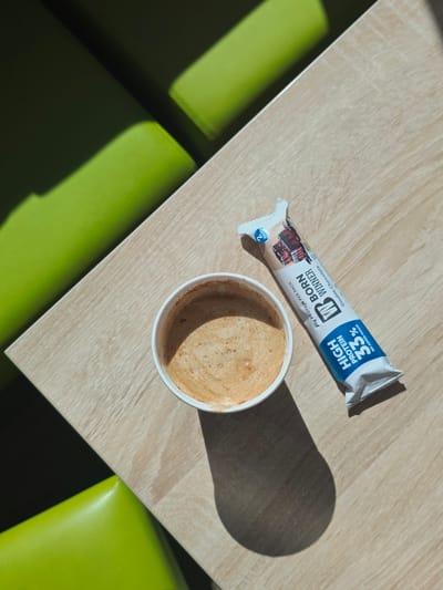An overhead, close-up shot shows a paper cup of coffee and a protein bar on a light wood grain table. The coffee has a foamy, tan-colored top. The protein bar wrapper is white with blue and red accents. The text on the wrapper reads "BORN WINNER," "Double Chocolate," "20g PROTEIN PER PACK," and "HIGH PROTEIN 35% NO ADDED SUGAR." The scene is lit with bright sunlight casting a large shadow of the coffee cup to the lower right. The background is blurred and shows out-of-focus green surfaces, suggesting a seating area with brightly colored furniture, possibly at a cafe or communal space in Drazhevo, Bulgaria. There are no people present.