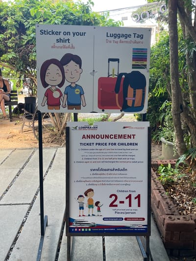 This image depicts a street-level scene with signage in front of lush greenery. The primary focus is on two vertical signs. The top sign, with a lighter background, is divided into two sections. The left section, titled "Sticker on your shirt," features illustrations of a smiling cartoon boy and girl wearing stickers on their shirts, with Thai text below. The right section, titled "Luggage Tag," shows illustrations of a red suitcase and a backpack, with a list of Thai destinations on colored tags.
The lower sign, with a white background and a dark border, is a "LOMPRA YAH HIGH SPEED CATAMARAN" announcement regarding "TICKET PRICE FOR CHILDREN." It lists conditions for children's fares, with the first condition in English and the second in Thai. Below this, there's a section in English titled "Children from 2-11" with text indicating "half price boat and car trips. Pieces/person." The sign also includes website addresses, "www.lomprayah.com" and "www.lomlahk.com," and social media icons (Facebook, Instagram, Line, WhatsApp) and contact information for customer service and online reservation.
In the background, a person is seated on a bench to the left, partially visible. The environment appears to be an outdoor area with trees and foliage, suggesting a location associated with travel or leisure, possibly a ferry terminal or a waiting area. The ground is paved with large square tiles interspersed with gravel. The overall setting suggests a tropical or subtropical climate given the dense vegetation. The image is taken from a medium-distance perspective, focusing on the informational signs.