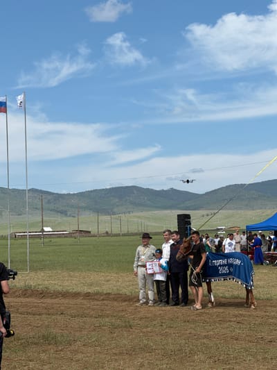 The image depicts an outdoor award ceremony with a horse at its center. A brown horse, adorned with a blue blanket featuring Cyrillic text "АЛТАРГАНА НААДАН 2026", stands to the right of the frame. A young man holds the horse's reins. To the left of the horse, several individuals are gathered. One man in a dark suit and another in a lighter suit stand beside a boy holding a certificate. A man in a brown hat stands to the far left of this group, also holding a certificate. Behind them, a crowd of spectators watches the proceedings. In the background, there are mountains, a large field, and several wooden structures that appear to be barns or outbuildings, suggesting a rural setting. A drone is visible flying in the sky above the ceremony. The sky is partly cloudy, with patches of blue. The ground is covered in grass and dirt. The presence of flags at the left of the frame indicates a formal event. The context suggests this is an award ceremony for a horse competition, possibly at a festival or cultural event in Kokorino, Russia.
