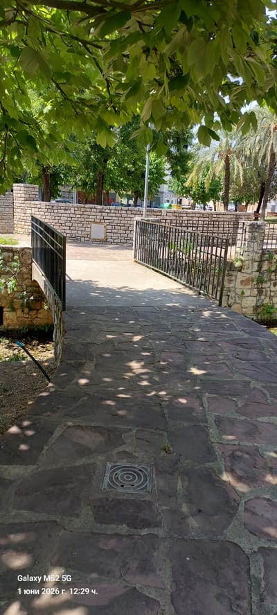 The image depicts a stone-paved area, likely a plaza or walkway, in the city of Canals, Spain. The pavement is irregular, with stones of varying sizes and shapes. Shadows from trees dappled the surface. Adjacent to the paved area are stone walls topped with metal railings, indicating a change in elevation or a boundary. Lush green trees and foliage are visible, suggesting a park or garden setting. A metal drain cover is embedded in the stone pavement. The date "1 юни 2026 г. 12:29 ч." is visible in the bottom right corner.