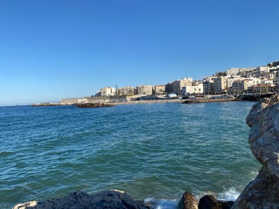 The image depicts a coastline in Beau Fraisier, Algeria. The foreground features the Mediterranean Sea with gentle waves, punctuated by dark, rocky outcrops. To the right, a large, light-colored rock formation partially obscures the view.
In the middle distance, a coastal cityscape stretches along the shoreline, consisting of closely packed, multi-story buildings, predominantly white or light-colored. The architecture suggests a mix of residential and commercial properties. A waterfront promenade runs along the base of the buildings, lined with trees and street furniture. There are people visible on the beach.
The sky is clear and bright blue, indicating sunny weather conditions. There are no boats visible on the water.