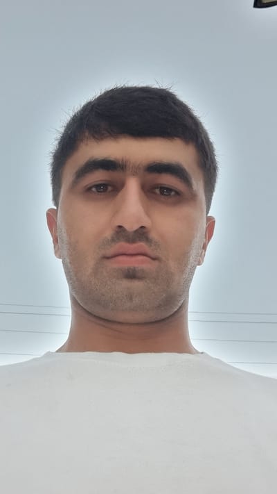 A close-up, low-angle selfie shows a man from the chest up, against a pale, overcast sky. The man has dark, short hair, thick eyebrows, and a light beard shadow. He is wearing a white t-shirt. In the background, faint power lines are visible against the sky. The image is a close-up, focusing on the man's face and upper body, and appears to have been taken outdoors. The location is the city of Khovaskor, Tajikistan.