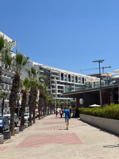 A sunny, clear day in Gżira, Malta, features a wide pedestrian walkway paved with light-colored bricks and decorative red rectangular inserts. Palm trees line the left side of the walkway, with their trunks secured with ropes. Beyond the palm trees, a multi-story, modern white building with balconies and dark-trimmed windows is visible. A few cars, including a blue one, are parked along the curb.
On the right side of the walkway, a contemporary building with dark wooden accents and large windows houses an establishment with umbrellas and outdoor seating. A man in a blue t-shirt and light shorts walks away from the camera down the center of the walkway. Other pedestrians are visible further down the path. In the background, construction cranes indicate ongoing development. The overall scene is one of a well-maintained urban environment with a resort-like feel.