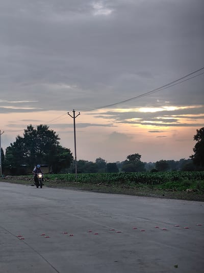 The image shows a paved road with a motorcyclist riding on it. The motorcyclist, wearing a plaid shirt and a helmet, is accompanied by a passenger also wearing a helmet. They are riding towards the viewer, with the motorcycle's headlight illuminated. In the background, there is a field of crops, trees, and a utility pole with electrical wires. The sky is cloudy with hints of sunset colors. The scene appears to be in Walsawangi, India, given the context provided. The road has reflective markers embedded in it, indicating it is a traffic zone. The overall atmosphere is calm and rural, with the natural light suggesting either dawn or dusk.