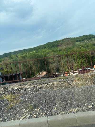 This image depicts a static, daylight scene in Tsarkva, Bulgaria, observed from a slightly elevated perspective. The upper third of the frame shows a pale blue sky partially covered by scattered, light grey stratiform clouds. Below the sky, a series of verdant hills covered in dense deciduous forest extends across the mid-horizon, displaying variations in green hues and foliage density.
In the midground, a prominent, dark brown, rusted metal fence with evenly spaced vertical bars runs horizontally, acting as a barrier. Behind this fence, several informal structures are visible amidst more sparse trees and vegetation. To the left, a building with a dark roof and a metallic satellite dish is present. Further right, behind the fence, additional structures with reddish-brown tiled roofs and other less defined architectural elements are discernible. Some trees directly behind the fence appear to have less foliage or are bare, contrasting with the lush hills.
The foreground consists of a coarse surface composed of dark grey and black gravel, interspersed with lighter-colored small stones and patches of dry, sparse vegetation and weeds. A light grey concrete curb with distinct segmentation lines runs along the very bottom edge of the image, indicating the boundary of a paved surface not fully visible within the frame. No individuals or active subjects are present in the scene.
