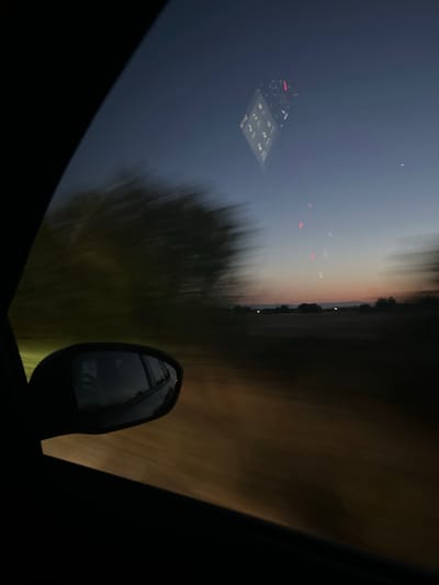 The image depicts a view from inside a moving vehicle at dusk or dawn. The foreground is dominated by the dark interior of the car, including the edge of the window frame and a side-view mirror. The mirror reflects a blurry image of the car's interior, including what appears to be a dashboard with illuminated gauges, suggesting the car is in motion.
The outdoor scenery is blurred due to the vehicle's movement, creating a sense of speed. On the left side of the image, blurred green foliage is visible, likely trees or bushes alongside the road. The sky transitions from a deep blue at the top to softer hues of orange and pink near the horizon, indicating either sunrise or sunset.
In the distance, a dimly lit landscape with silhouetted trees and possibly some faint lights from structures can be observed. There are also a few blurry red and white lights visible in the sky, which could be aircraft lights or other atmospheric phenomena.
The location is described as Skutare, Bulgaria. This implies the scene is set in or around this Bulgarian locality. The overall mood is serene and transient, capturing a moment of travel during twilight.