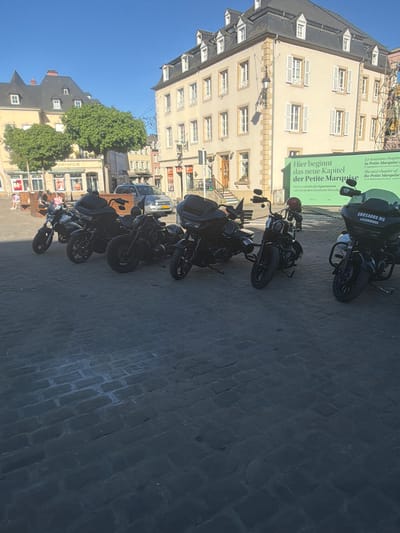 The image shows a street in Echternach, Luxembourg, with several motorcycles parked along the side of the road. A building with a sign written in both German and French is visible in the background, translating to "The new chapter of the Petite Marquise." There are also a few people visible in the image, walking or sitting on benches near the motorcycles. The sky is clear and blue, indicating it is daytime. The street is paved with cobblestones.