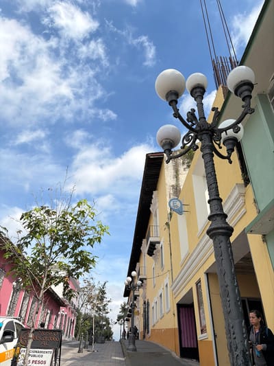 A street scene with a vintage lamppost in the foreground. The lamppost has five globe lights and ornate metalwork. Behind, a row of yellow buildings lines the street, some with awnings. A person peers out of an upper window. On the right, pink buildings and trees are visible, along with a sign that appears to be for a museum. A blue sky with scattered clouds fills much of the frame.