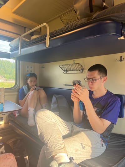 The image shows two young people inside a train compartment. The compartment appears to be a sleeper car, with a blue upholstered bench seat that can be converted into a bed. The lighting suggests it is daytime, with sunlight streaming in through the train window, casting shadows and highlighting the scene. The overall mood is relaxed and casual, with the two individuals engaged in their own activities.
On the right side of the frame, a young man wearing glasses, a purple t-shirt, and gray sweatpants is holding a smartphone and appears to be taking a picture or video of himself or his surroundings. He is seated on the bench and his legs are extended. Visible text on his t-shirt includes "SPORT" and possibly "RUSSIA". The sweatpants have a "PUMA" logo.
On the left side of the frame, a young woman is curled up on the bench, also looking at a smartphone. She is wearing a blue and white top, white socks with a black logo, and sunglasses perched on her head. She is facing the window, with her knees drawn up to her chest.
The train compartment is furnished with the standard amenities, including a small table, a metal rack attached to the wall, and coat hooks. A window on the left shows a glimpse of green trees and a building, suggesting the train is moving through a rural or suburban area. The image is taken from a slightly elevated, tilted perspective, adding to the candid feel of the shot. The location context provided is Пугачёво, Russia.