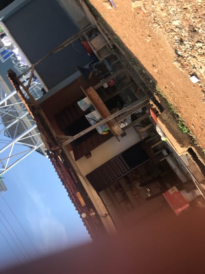 The image, taken at an angle, depicts an outdoor scene in what appears to be a residential or commercial area within Mandate Estate, Ilorin, Nigeria. The foreground shows a sun-drenched, unpaved dirt path or road covered with loose soil, small rocks, and debris. Patches of green vegetation are visible along the edge of this path.
To the right, a multi-level structure or building dominates the view. It features wooden beams and supports, suggesting ongoing construction, a makeshift addition, or a weathered existing structure. Visible elements within and around the building include dark brown doors, one with horizontal slats, and multiple windows, some with white frames. Various items are stored or displayed within the structure, including white plastic containers or bags and colorful fabrics, one with red polka dots and another with a white and green pattern. A prominent red box with white lettering is visible, clearly showing the word "KIMCHI" and other text such as "PRESERVED CABBAGE." A corrugated metal roof extends over parts of the structure, and a light fixture is attached underneath. In the lower part of the image, a metal framework or scaffolding is visible against the sky.
The background reveals a clear, bright blue sky with a few faint wispy clouds, indicating sunny weather. Utility wires stretch across the sky.
No people are visible in the image, and no specific activity is taking place; the scene appears static. The bright sunlight and sharp shadows suggest it is daytime, likely mid-morning or afternoon. The overall setting, with unpaved roads and the type of building construction, aligns with the context of a developing area.