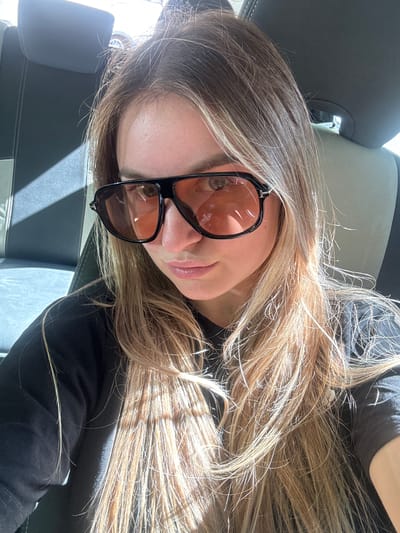 A young woman with long, blonde hair and sunglasses is shown from the chest up, in a car. The interior of the car is visible behind her, with a black and grey seat and headrest. Sunlight streams in from the right, casting shadows across her hair and face. She is wearing a black t-shirt. The specific location in L'Essard, France is not discernible from the image.