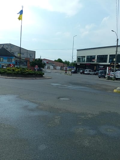 The image captures a street scene in Tiachiv, Ukraine, characterized by a roundabout in the foreground with wet asphalt, indicating recent rain. A prominent Ukrainian flag on a tall pole flies to the left, silhouetted against a pale blue sky with scattered clouds. To the left of the roundabout, a multi-story building with visible windows and air conditioning units is present. Adjacent to it, a smaller structure with a dark roof and blue and white signage is partially visible.
Further into the scene, a modern commercial building with large windows and a white facade stands to the right. Signs on this building include "FIMKA", "FASHION SHOPPING", and "Neo-bile". Several vehicles are parked along the street in front of this building, including a white van, a dark sedan, a dark minivan, and another white vehicle. A traffic light and street lamps are also visible. In the background, other buildings and trees are present, suggesting an urban or semi-urban environment. Traffic signs are visible, including directional signs with blue backgrounds and white text, as well as red and white chevron directional signs around the roundabout. The overall impression is a moderately busy street on a cloudy day.